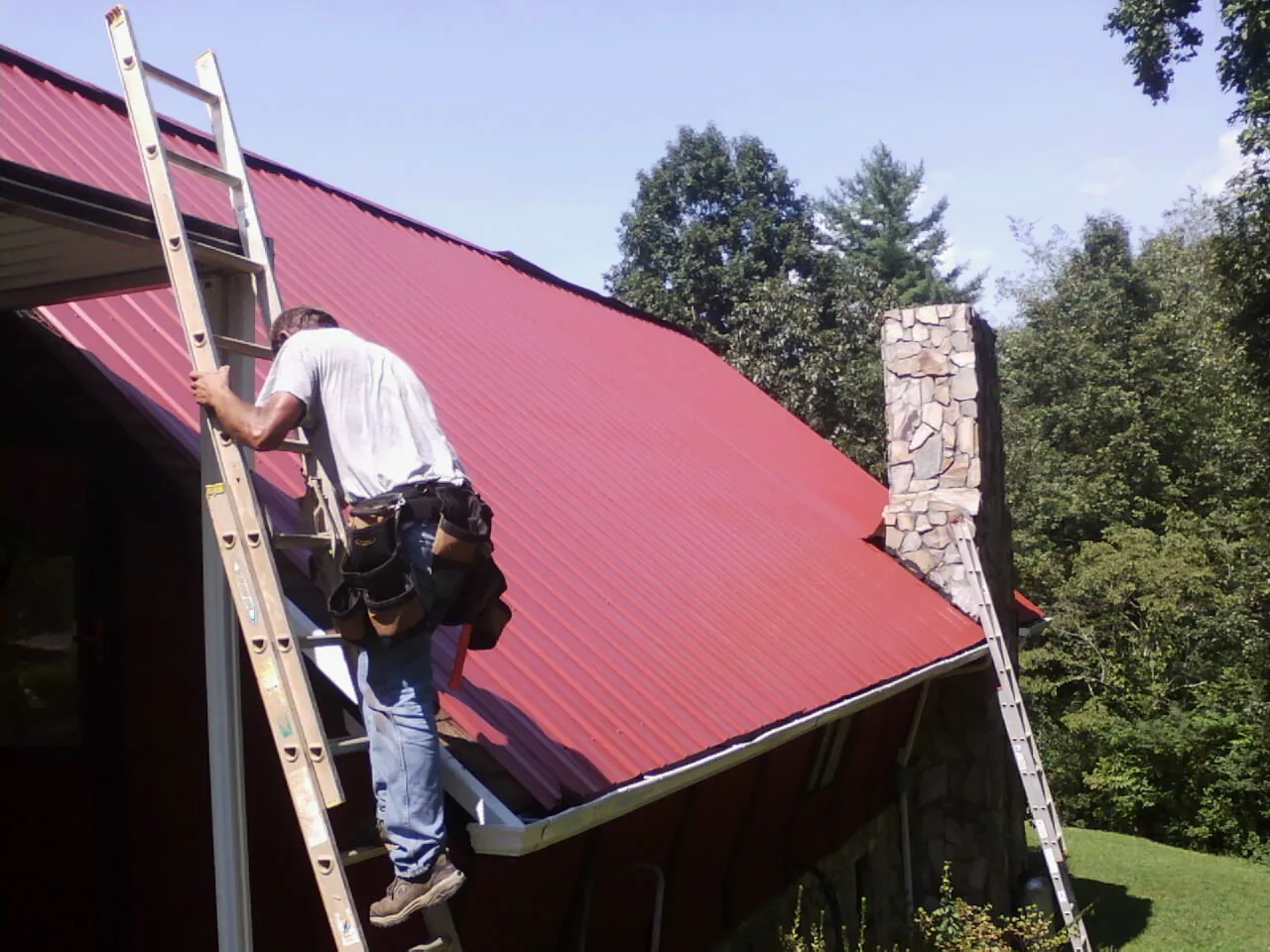 Roofer on a ladder beside a red metal roof and stone chimney — Soffit & Fascia Repair by Roofing Brothers in Alexis
